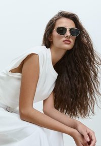 Occhiali da sole aviator con lenti scure e montatura in metallo argentato. Modello che indossa una maglietta bianca con maniche a taglio, in posa contro uno sfondo neutro.
