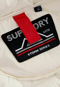 Krämfärgad tyg med en svart lapp som visar "SUPPORT DRY MTN" och "STORM SERIES," med ett rött accentband och grå designelement.