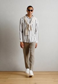 Gestreiftes Hemd mit langen Ärmeln, hellbeige Chinos, weiße Sneaker und ein cremefarbener Pullover, der über die Schultern gelegt ist.