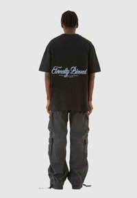 Tricou din bumbac negru, cu mâneci scurte și croială lejeră, având textul albastru "Eternally Blessed" pe spate. Purtați cu pantaloni cargo închiși la culoare.