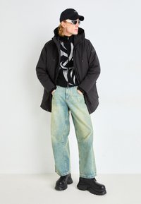 Giacca nera con cappuccio e zip, maglione grafico bianco e nero, jeans svasati blu chiaro e stivali neri massicci, completati da un berretto nero e occhiali da sole.