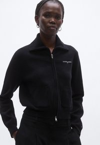 Fekete, zipzáras dzseki gallérral, két első zsebbel, bordázott mandzsettákkal és szegéllyel; fehér hímzett logóval a bal mellrészen.