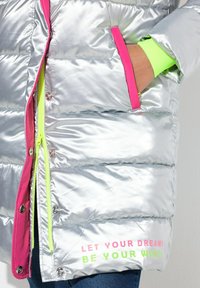 Doudoune argentée avec des accents roses, fermeture éclair vert fluo et texte "LAISSEZ VOS RÊVES ÊTRE VOS AILES !" sur l'ourlet. Comprend des poches latérales.