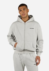 Sudadera gris con cremallera, bolsillo canguro, puños y dobladillo acanalados, presenta un logo negro de "PEGADOR" en el pecho. Material suave de mezcla de algodón.
