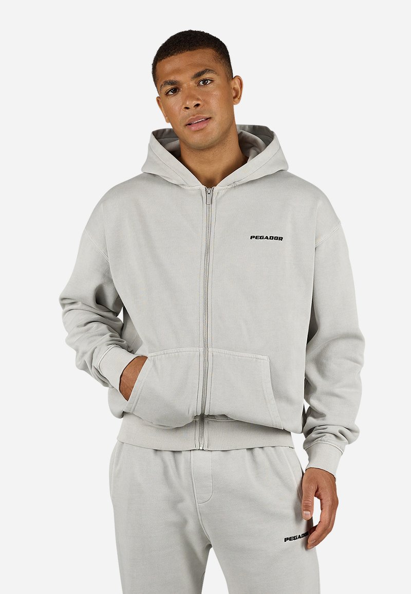 Sudadera gris con cremallera, bolsillo canguro, puños y dobladillo acanalados, presenta un logo negro de "PEGADOR" en el pecho. Material suave de mezcla de algodón.