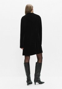 Schwarzes Samtkleid mit langen Ärmeln und hohem Ausschnitt. Das Design hat eine gerade Silhouette und einen Reißverschluss auf der Rückseite. Getragen mit kniehohen schwarzen Stiefeln.