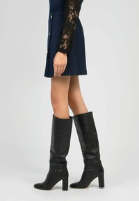 Femme portant des bottes en cuir noir jusqu'aux genoux avec des talons blocs, une jupe marine à boutons et un haut en dentelle noir à manches longues.