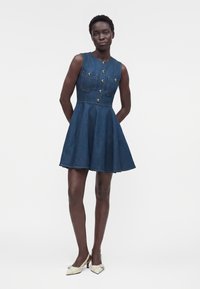 Femme portant une robe en jean bleue sans manches avec des boutons dorés et des poches avant, associée à des chaussures crème à petit talon, debout les mains dans le dos.
