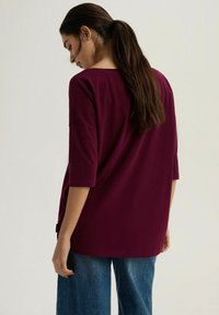 Burgundowy luźny top z rękawami 3/4, z okrągłym dekoltem i bocznymi rozcięciami, w połączeniu z luznymi niebieskimi dżinsami.