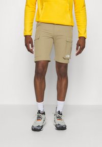 Man i beige cargo shorts, vita strumpor, grå och svarta vandringsskor samt en gul sweatshirt mot en neutral bakgrund.