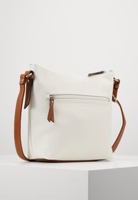 Baltā ādas crossbody soma ar brūno siksnu, ar rāvējslēdzēja kabatu un dekoratīviem rāvējslēdzēja tagiem, maiga tekstūra un strukturēta forma.