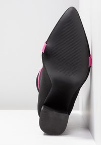 Mustat neulekorkokengät, joissa on teräväkärkinen muoto, vaaleanpunainen korostushihna ja teksturoitu pohja, esiteltynä takaa.