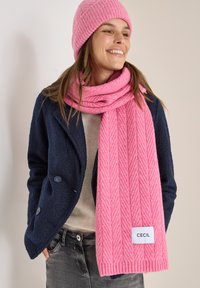 Rosa gestrickter Schal mit einem Fischgrätmuster und gerippten Kanten, kombiniert mit einer passenden Mütze, getragen über einem marineblauen Mantel und einem grauen Oberteil.