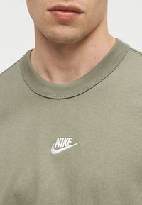 Κοντινό πλάνο ενός ατόμου που φοράει μπλούζα Nike ελιάς με λευκό κεντητό λογότυπο στο κέντρο του στήθους.