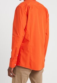Man in een feloranje lange mouwen overhemd met knopen, gecombineerd met beige broek, gezien van achteren tegen een witte achtergrond.