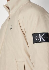 Beżowa kurtka zapinana na zamek z wysokim kołnierzem, małym logo "ck" z przodu oraz czarną naszywką na rękawie z napisem "Calvin Klein Jeans cK."