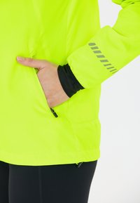 Bright lime green windbreaker with a pocket and reflective gray accents. Fabric is smooth, lightweight, with a relaxed fit. Black base layer visible.