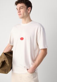 Hvid bomulds t-shirt med rund halsudskæring, korte ærmer og en rød logoaccents i midten. Modellen holder en brun stofpose.