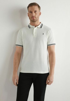 Mężczyzna w białej koszulce polo z krótkim rękawem, z czarnymi paskami na kołnierzyku i rękawach, stojący na tle jednolitej białej ściany.