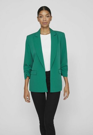 Grüner Blazer mit lässiger Passform, mit Revers und hochgerollten Ärmeln. Getragen über einem weißen Oberteil und kombiniert mit schwarzen Hosen. Glatte Textur.