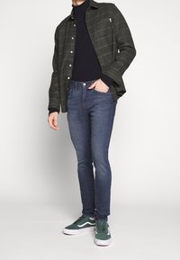 Mörkgrön rutig skjorta, marinblå polotröja, blå slim-fit jeans och gröna mockasneakers med vita accenter. Texturerad tyg, avslappnad design.