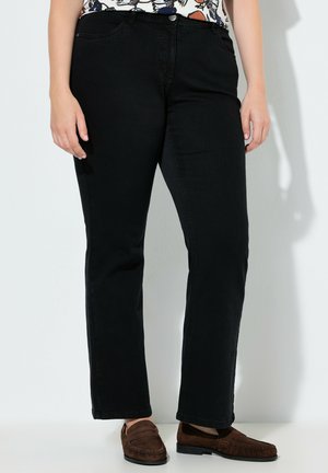 Person iført sorte jeans med lige ben, brune loafers i ruskind og en mønstret top, stående op ad en hvid væg.