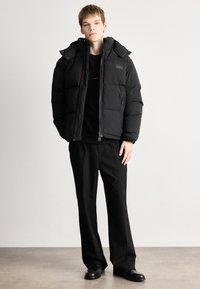 Crna zimska jakna s kapuljačom, s prednjim zatvaračem i bočnim džepovima, nosi se preko crne dukserice. Uparena je s crnim hlačama i cipelama.