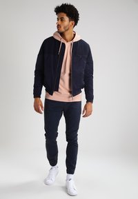 Giubbotto bomber navy su un hoodie rosa chiaro, abbinato a pantaloni in denim scuro e sneakers bianche. Il giubbotto ha una chiusura con zip e tasche.