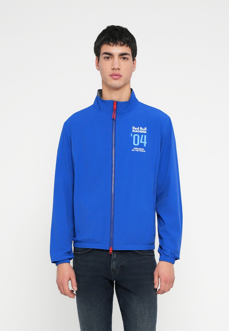 Blauwe lichte jas met een hoge kraag, ritssluiting aan de voorkant en een contrasterende rode ritslip. Voorzien van een gedrukt logo op de borst.