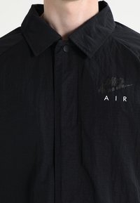 Černá nylonová bunda s skládacím límcem, zapínáním na patentky a reflexním logem Nike na levé straně. Má texturovaný povrch.