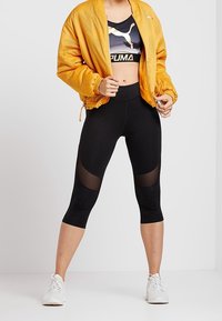 Czarne legginsy capri z bocznymi panelami z siateczki, w połączeniu z krótka czarną i złotą bluzą sportową oraz jaskrawą pomarańczową bomberką z siateczki.