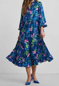 Květinové šaty ve stylu A-line v modré, zelené a růžové barvě, s rukávy 3/4, výstřihem do V a volánkovým lemem. Kombinované s modrými vysokými podpatky.