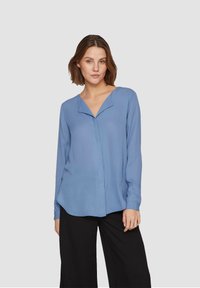 Blusa azzurro chiaro a maniche lunghe con scollatura a V e un sottile plissè frontale, abbinata a pantaloni neri a gamba larga. Tessuto morbido e fluente.