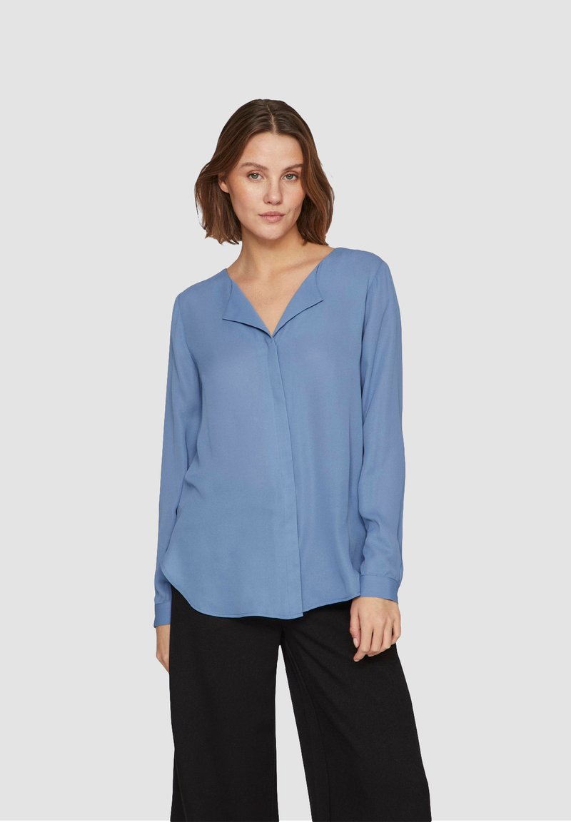 Blusa azzurro chiaro a maniche lunghe con scollatura a V e un sottile plissè frontale, abbinata a pantaloni neri a gamba larga. Tessuto morbido e fluente.