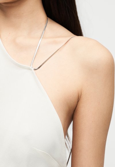 Lähivaade õlast, mis kannab valget sümmeetrilist topi õhukese metallist kettidega rihmaga ja mille taga on nähtav pikk tume juukse.