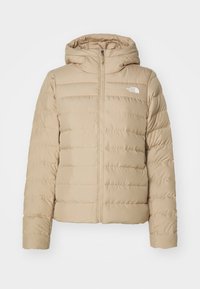 Doudoune beige avec capuche, fermeture à glissière sur le devant et panneaux cousus. Un petit logo blanc est présent sur le côté gauche de la poitrine. Texture de tissu douce.