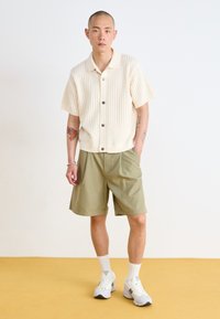 Beige knit short-sleeve cardigan with buttons, paired with olive green shorts. Model wears white socks and gray running shoes, standing on a yellow floor.