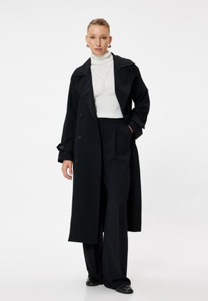 Black trench coat with large lapels, double-breasted buttons, and side pockets. Worn over a white turtleneck and black wide-leg trousers.