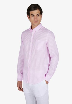 Homme portant une chemise à manches longues rayée rose clair avec un pantalon blanc, debout, une main dans la poche, regardant vers la gauche.