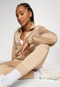 Beige träningsoverall med dragkedja och ribbade mudd, kombinerad med ljusa sneakers och vita strumpor, visad i en sittande pose.