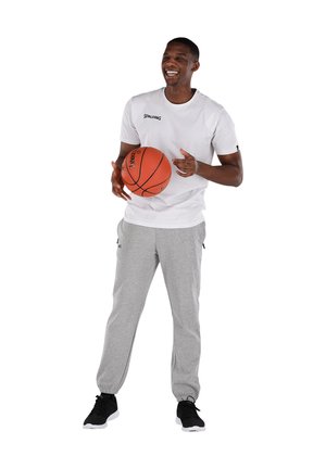 Witte T-shirt met korte mouwen met het "SPALDING" logo, grijze joggingbroek met zijzakken, zwarte sneakers, een oranje basketbal vasthoudend.