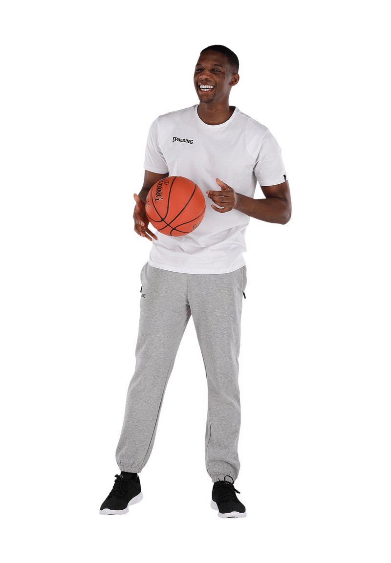 Witte T-shirt met korte mouwen met het "SPALDING" logo, grijze joggingbroek met zijzakken, zwarte sneakers, een oranje basketbal vasthoudend.