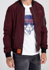 Giacca bomber color borgogna con una texture liscia e lucida, polsini a coste e una tasca con zip sulla manica. Presenta un'etichetta arancione del marchio sull'orlo.