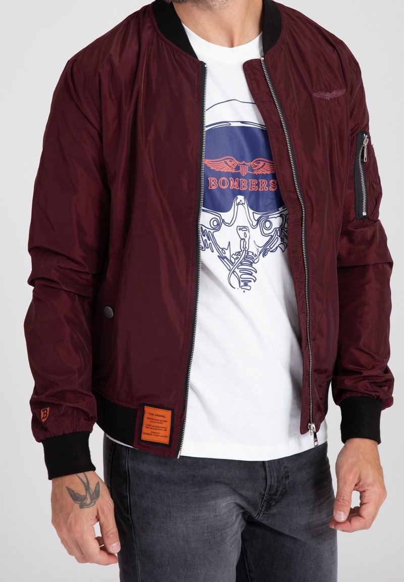 Giacca bomber color borgogna con una texture liscia e lucida, polsini a coste e una tasca con zip sulla manica. Presenta un'etichetta arancione del marchio sull'orlo.