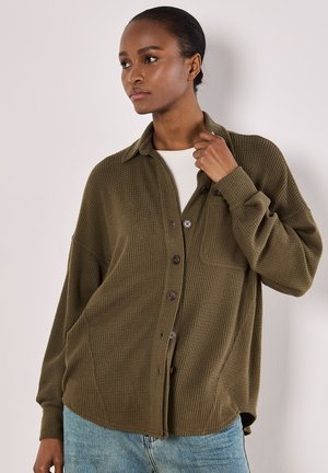 Camisa de botón texturizada en verde oliva con cuello, que cuenta con un bolsillo en el pecho y un corte relajado. El modelo la lleva puesta sobre una camiseta blanca y unos jeans de mezclilla.