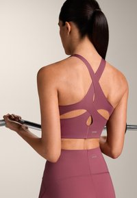 Donna che tiene un bilanciere, indossa un reggiseno sportivo malva con spalline incrociate e leggings a vita alta, vista di spalle.