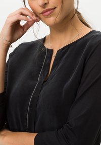 Musta paita, jossa on pyöreä kaula-aukko ja edessä nappilista. Pitkät hihat, joissa on teksturoitu pinta. Hopeaketju, jossa on pieni riipus.