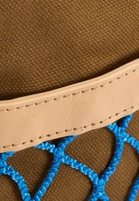 Καφέ υφασμάτινο φόντο με υφή, μπεζ δερμάτινο λουράκι με ραφές και μπλε πλεγμένο κορδόνι που σχηματίζει μοτίβο δίχτυ κάτω από το λουράκι.