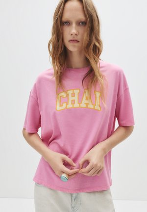 Rosa T-Shirt mit kurzen Ärmeln, das mit auffälligen gelben "CHA!"-Buchstaben auf der Vorderseite versehen ist. Weiches Material mit entspannter Passform.