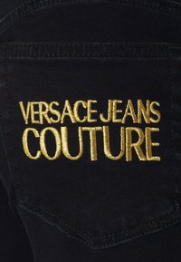 Gold embroidered "Versace Jeans Couture" logo on the back pocket of dark denim jeans with visible stitching.